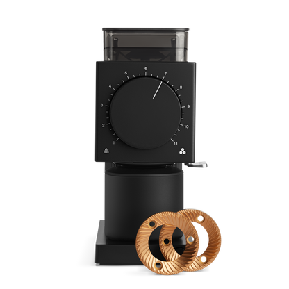 The Fellow Wholesale Ode Brew Grinder, featuring a sleek black design with a square top and a large circular dial in the center, is displayed. Beside the grinder, two golden Gen 2 Brew Burrs are showcased, highlighting its innovative components. Set against a black background, the grinder and burrs stand out with their impressive 31 grind settings and anti-static technology.