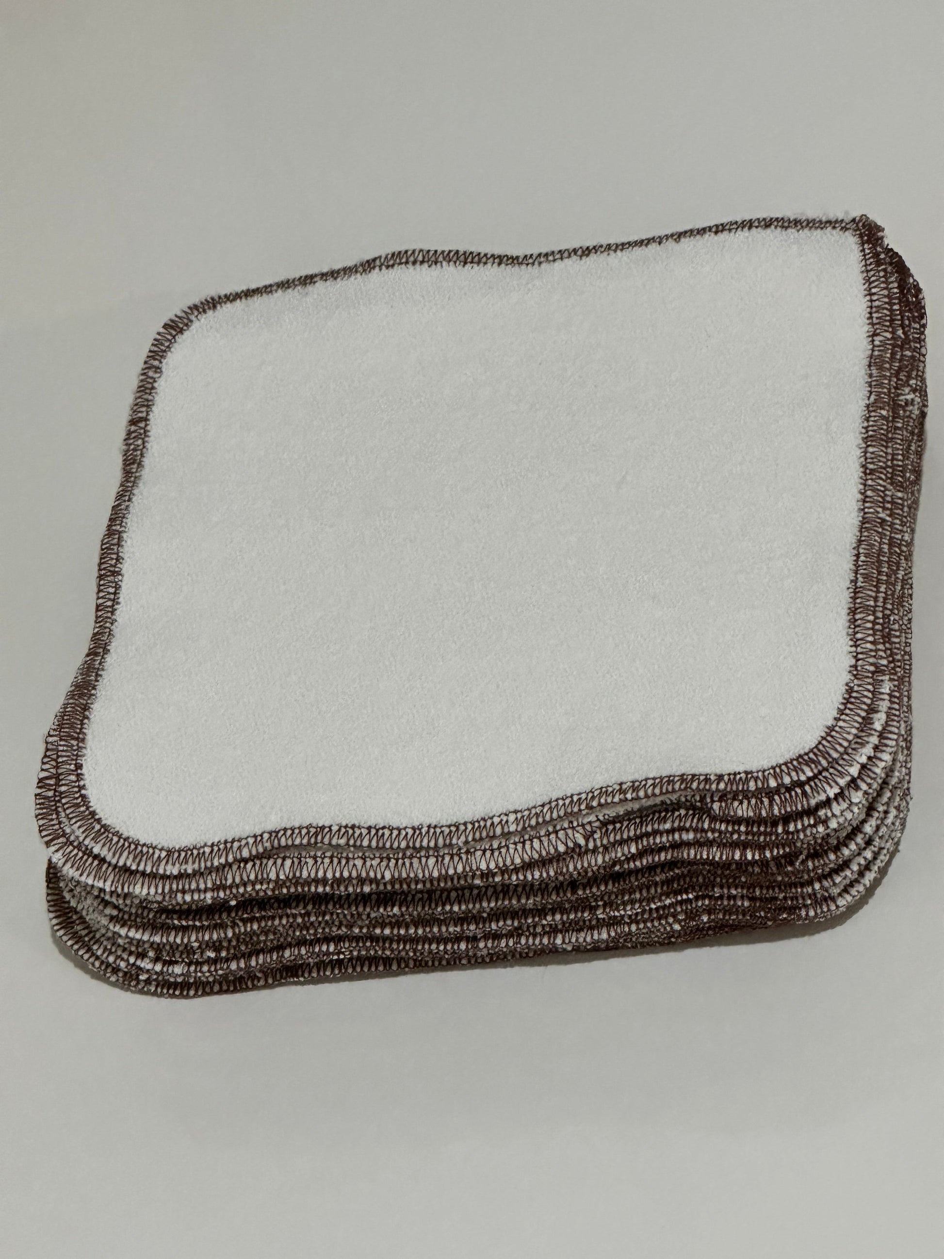 The ORGANIC WASHCLOTH in cocoa by cabane childrenswear features a stack of white square cloths with brown stitched edges, handmade from organic cotton in Vancouver. Soft and absorbent, they are ideal for cleaning or personal use.