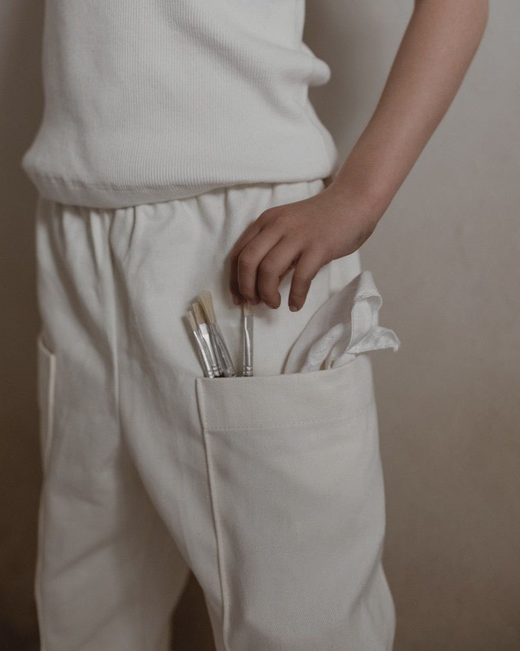 A person dons the Pantalon D'Artiste in Hemp Twill by cabane childrenswear, showcasing their white loose-fitting pants. Their hand rests on a pocket brimming with paintbrushes and a cloth against a neutral background that complements this ethically handmade, creativity-evoking ensemble.