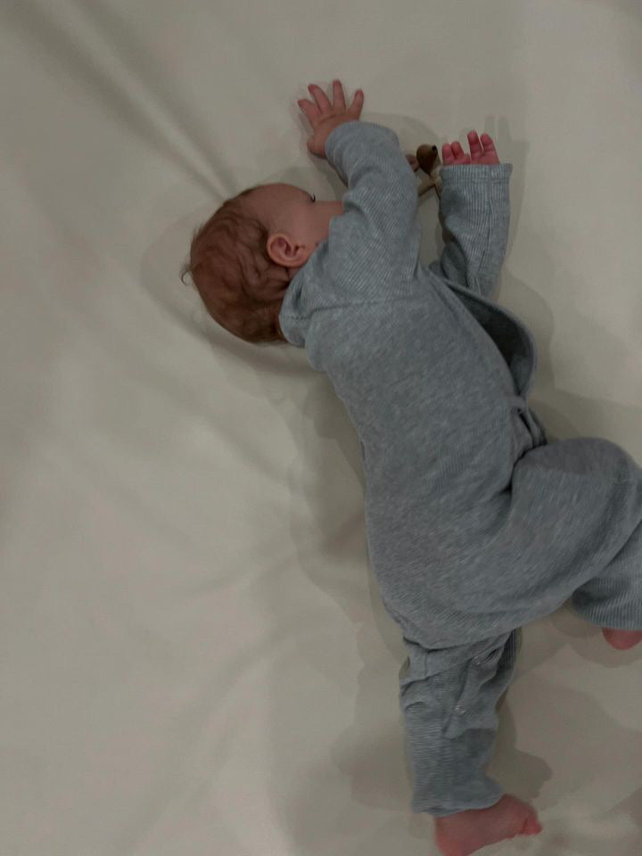 A baby in the gray APRÈS WRAP organic waffle onesie from cabane childrenswear lies on a light-colored sheet. The baby is turned to the side with arms stretched out and legs bent, enjoying the softness of ethically handmade clothing.