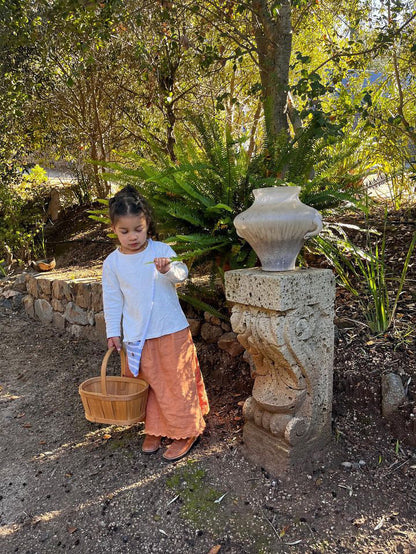 A young girl with braided hair stands in a garden holding an empty wicker basket. Dressed in the cabane childrenswear RESORT TOP hemp jersey and a peach skirt, she touches a stone pedestal with a large decorative vase on top. The garden, lush with greenery and sunlight, offers the perfect backdrop for her handmade attire crafted in Calgary.