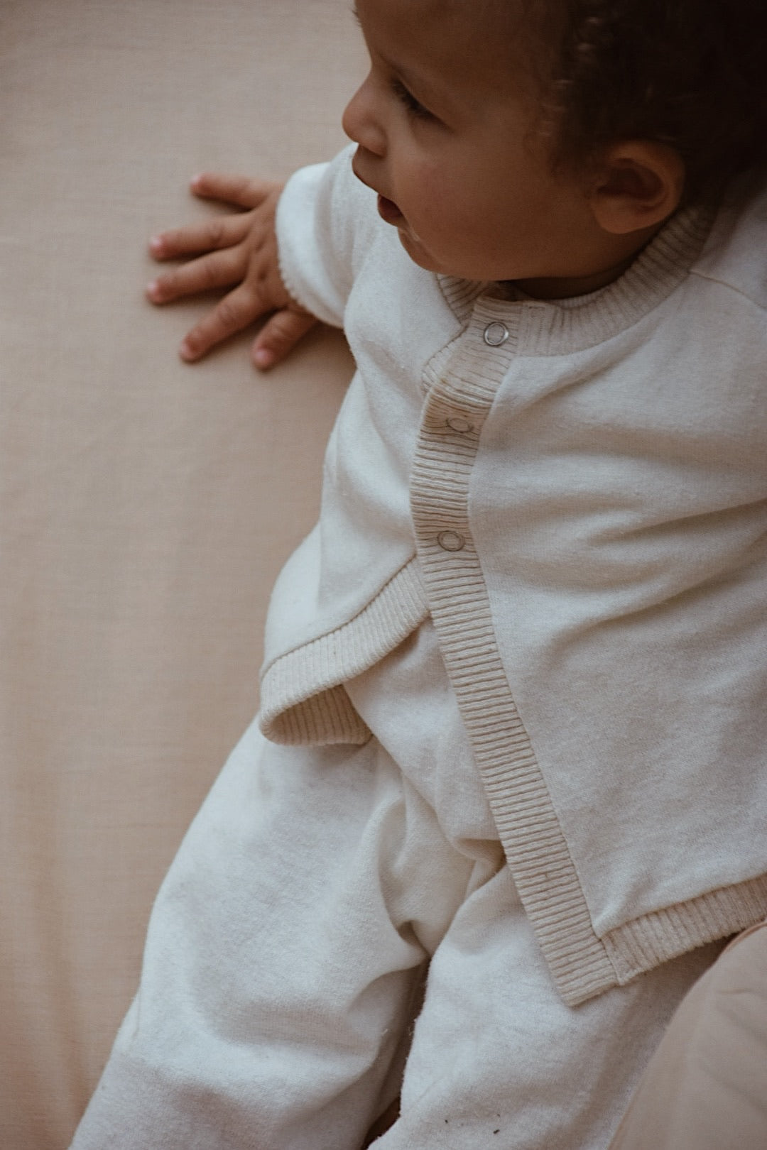 A young child wearing the ethically handmade, cozy Soul Sweater Baby + Kid from cabane childrenswear, featuring a boxy 3-snap sweater in organic cotton and hemp, sits on a soft beige surface. The child looks to the side with one hand resting on the surface for support.