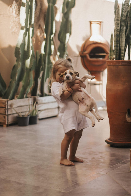 A young child in a pre-order HIGH-NOON SUNSUIT by cabane childrenswear joyfully holds a small dog while standing on a tiled patio. Surrounding them are large potted plants and cacti, with a terracotta chiminea in the background. The scene is warm and sunlit, creating a cozy, inviting atmosphere.