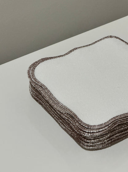 A stack of ORGANIC WASHCLOTHS by cabane childrenswear, featuring a white square design with brown edges made from hemp french terry, rests on a white surface. Their textured look and neat stitching stand out against a plain light-colored wall, exuding elegance and quality.