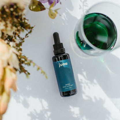 A bottle of Juna Chlorophyll Detox Drops, an herbal dietary supplement promoting liver detoxification, sits on a white surface next to a glass of green liquid. Some blurred flowers are partially visible on the left side of the image.