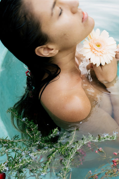 A person with closed eyes relaxes in the Surya Love Bath Heart Opening Soak, partially submerged. They hold a light pink flower close to their face. Their dark hair is wet and their shoulders are bare. Surrounding them are green plants and rose petals floating gracefully on the water.