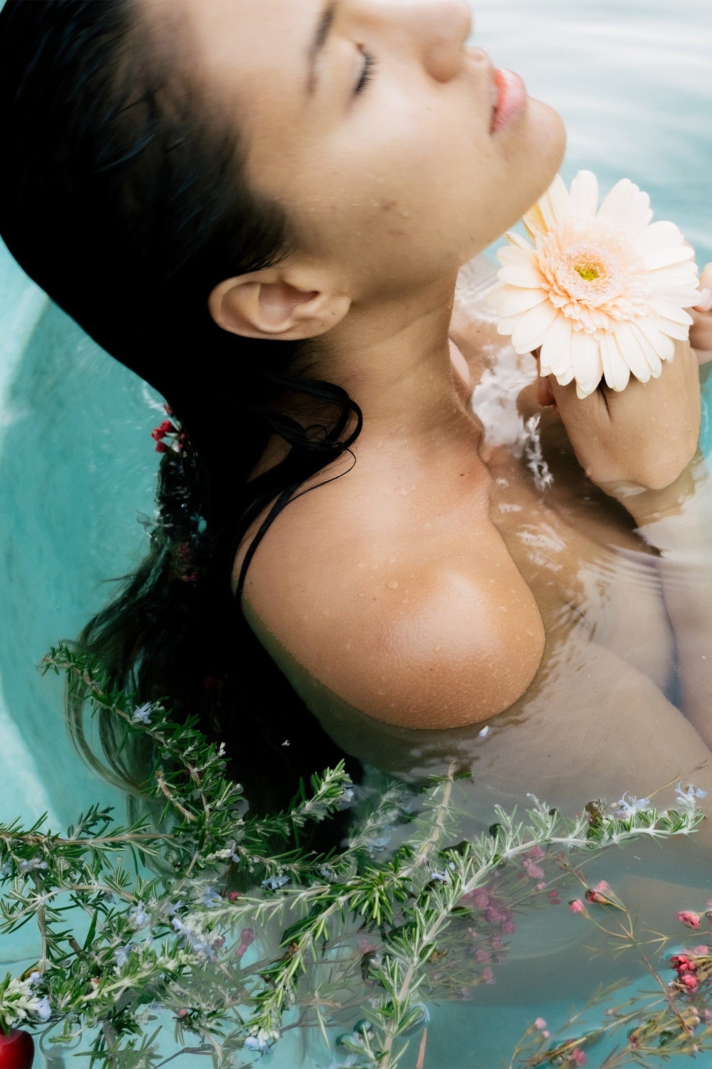 A person with dark hair is enjoying a calming bath soak, eyes closed and head tilted back while using Surya's Cooling Bath Soak. They hold a pale orange flower near their shoulder, surrounded by lush greenery and water. The scene is both serene and relaxing.