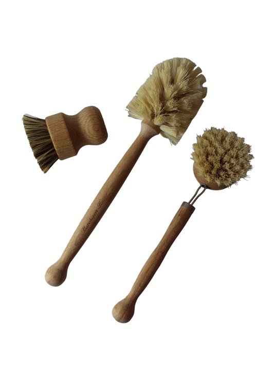 The All-Brush Bundle by Murchison-Hume is displayed on a white background, featuring three wooden cleaning brushes with natural fiber bristles. The set includes a short mushroom-shaped brush, a long-handled brush with an angled head, and a round brush with a slightly bent handle. These compostable brushes are ideal for eco-friendly cleaning.
