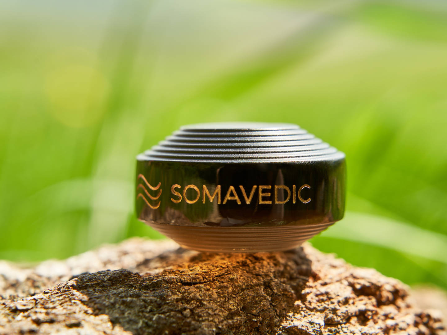 A close-up shot of a compact, dome-shaped device from Somavedic USA with the brand name "SOMAVEDIC" engraved on it, placed on rough tree bark. The background is a soft blur of green and yellow, evoking an outdoor environment with natural lighting. This charged Tiny device is renowned for creating a protective field.