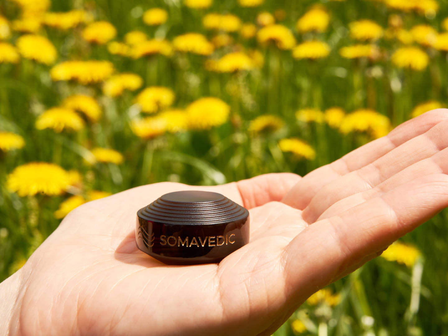 A hand holding a compact, dark-colored device named "Tiny" by Somavedic USA with the brand name engraved on it. The background is a field of bright yellow flowers in focus. This charged device appears to be smooth and creates a protective field around it.