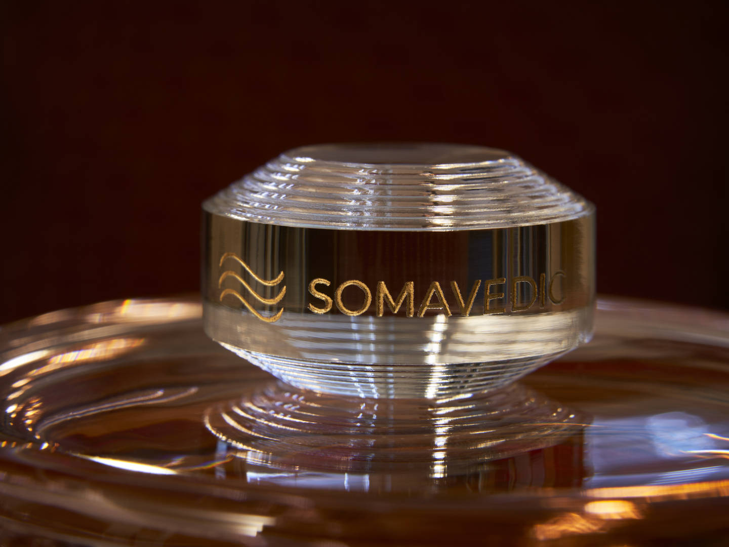 Close-up image of a clear, circular glass top with ridged texture and metallic gold accents. The top, part of the Tiny device by Somavedic USA, features the engraved text "SOMAVEDIC" and a wavy line graphic. The background is blurred and dark, highlighting the clarity and reflections of the glass's protective field.