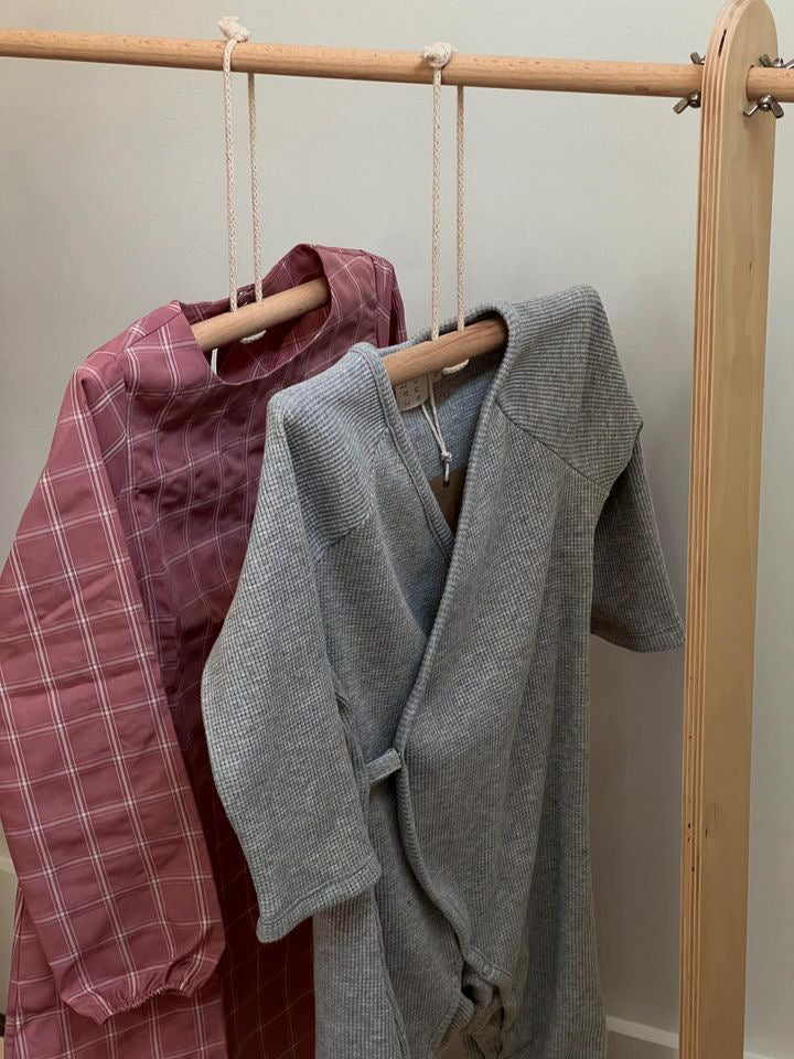 Two baby outfits hang on a wooden rack, each crafted from soft organic cotton. One is a red plaid dress, while the other is the APRÈS WRAP organic waffle, a grey wrap-style onesie by cabane childrenswear. Both garments are specially designed for newborns with care and ethics in mind and are suspended by string-attached hangers.