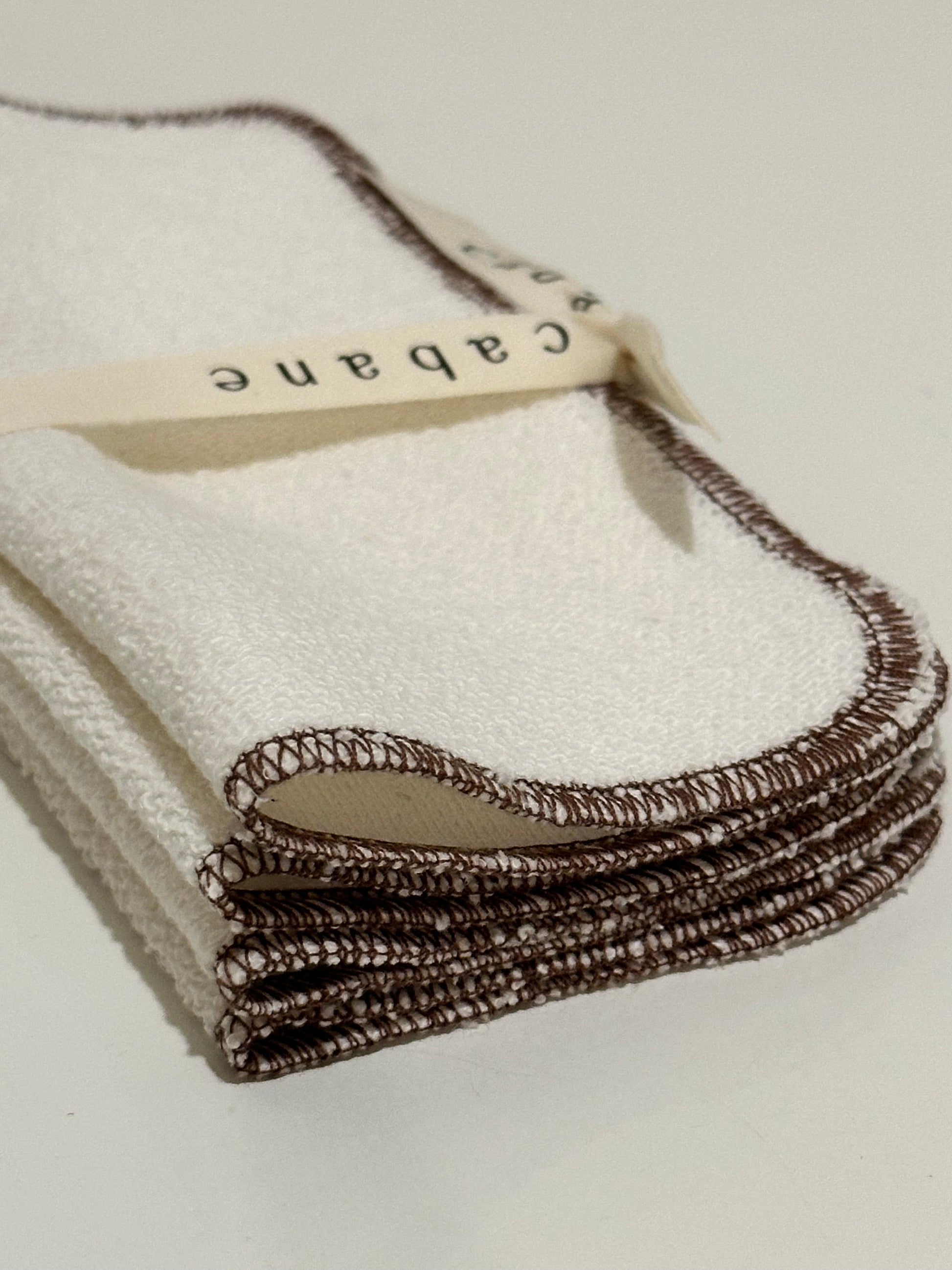 A stack of "cabane childrenswear" ORGANIC WASHCLOTHs in cocoa, handcrafted in Vancouver, are neatly folded with dark brown edges and tied with a ribbon labeled "cabane," displayed on a plain, light surface.