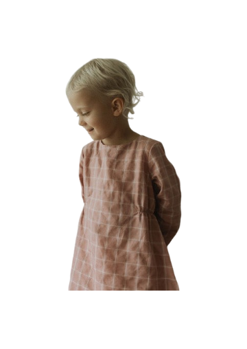 A young child with short, blond hair stands turned sideways, looking downward with a gentle smile while wearing the DAY DRESS plaid by cabane childrenswear. This limited edition print in pink raspberry exudes charm as their hands rest behind their back against a simple white backdrop.