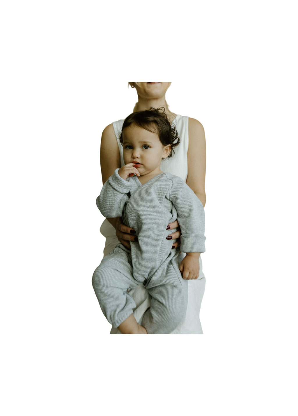 A baby wearing the APRÈS WRAP organic waffle outfit by cabane childrenswear is cradled by an adult in a white dress. The baby's dark hair frames their face as they softly suck on a finger and gaze at the camera. The plain, dark background enhances the tranquility of the scene, while the adult's face stays hidden from view.