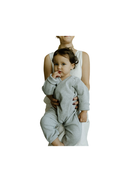 A baby wearing the APRÈS WRAP organic waffle outfit by cabane childrenswear is cradled by an adult in a white dress. The baby's dark hair frames their face as they softly suck on a finger and gaze at the camera. The plain, dark background enhances the tranquility of the scene, while the adult's face stays hidden from view.