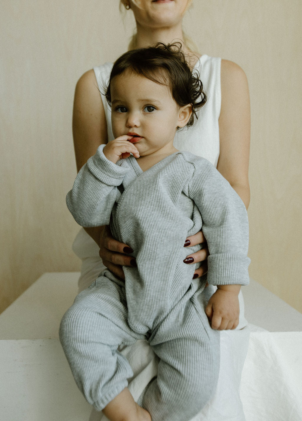 A woman in a white dress cradles a young child dressed in an APRÈS WRAP organic waffle outfit by cabane childrenswear. The child, nestled on her lap, looks at the camera with fingers in their mouth against a simple, light-colored wall.