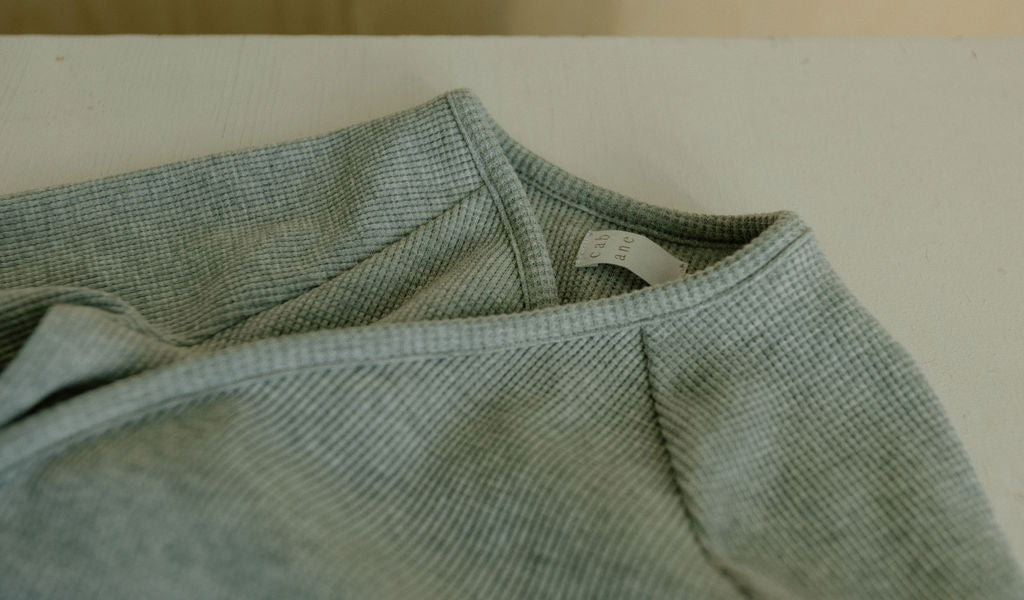 A close-up of a light green textured fabric garment, the APRÈS WRAP organic waffle by cabane childrenswear, rests on a light surface. A small white tag with printed text is partially visible at the neckline.