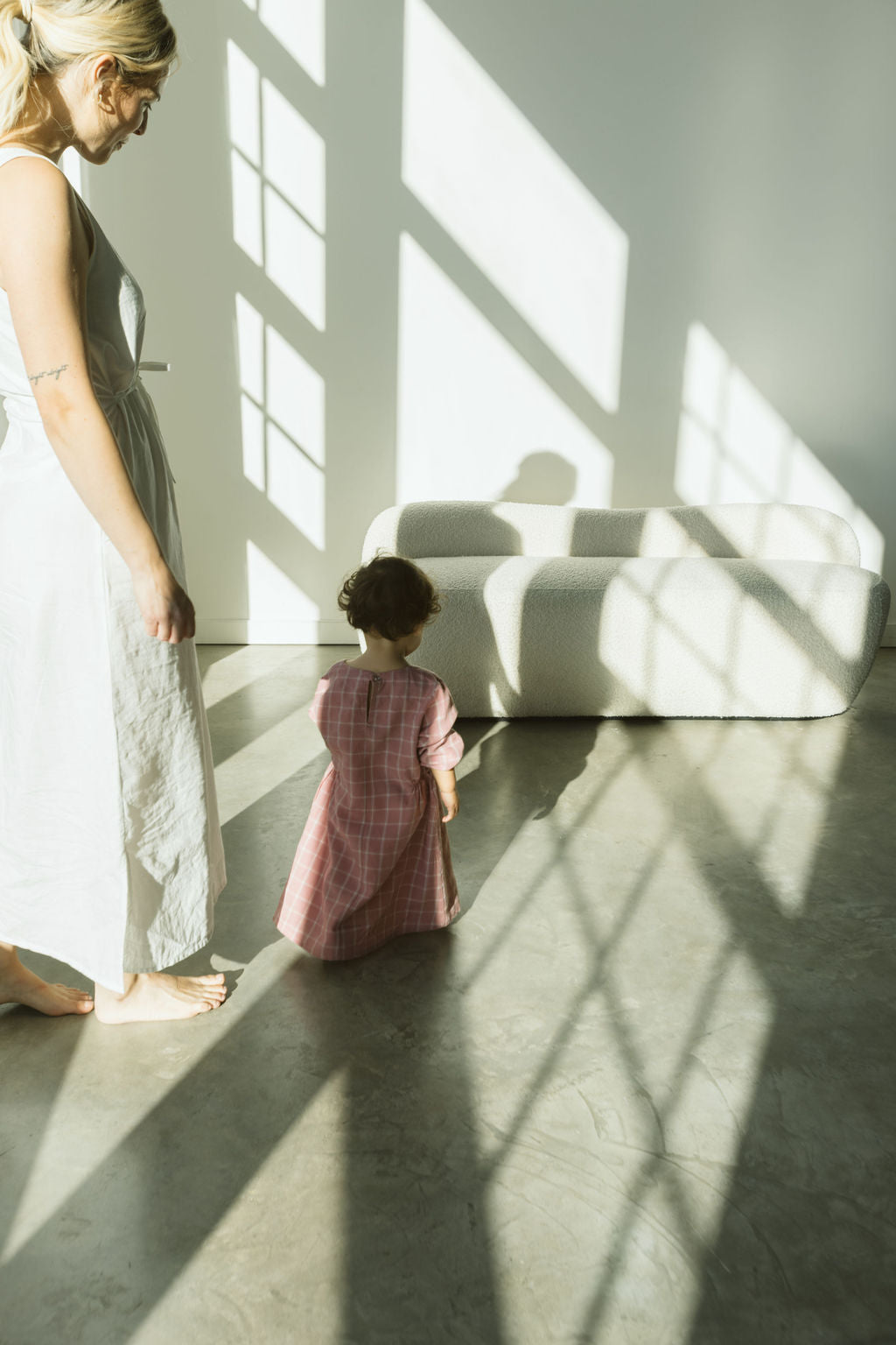 A woman in a white dress and a toddler wearing the DAY DRESS plaid from cabane childrenswear stroll across a sunlit, polished concrete floor. The shadows of window panes form a charming grid pattern, enhancing the scene's appeal. In the background, a limited edition white sofa completes this serene tableau.