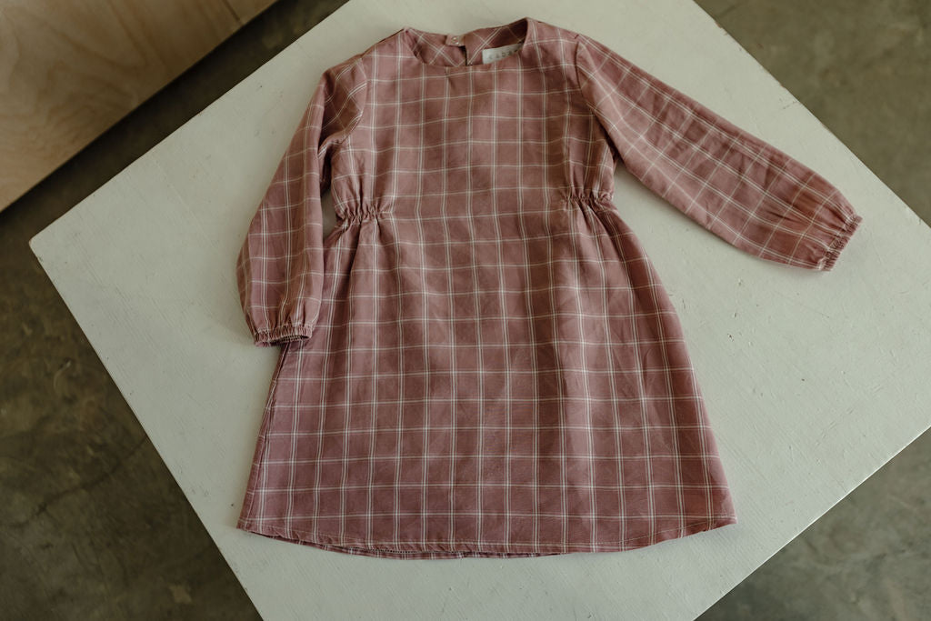 Displayed on a white surface, the DAY DRESS plaid by cabane childrenswear features a long-sleeved, pink plaid design with elegantly gathered sides at the waist. Crafted from exclusive raspberry plaid fabric, this dress is a limited edition piece handmade in Vancouver.