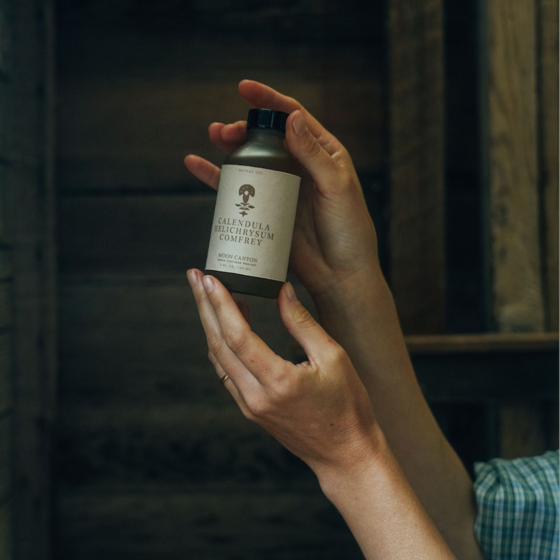 A person holds a small brown bottle labeled "Calendula, Helichrysum & Comfrey Herbal Oil" with both hands in front of a wooden backdrop. The bottle features an image of a tree and some text underneath the primary label from Moon Canyon Healing. The person appears to be examining the face and body oil closely.