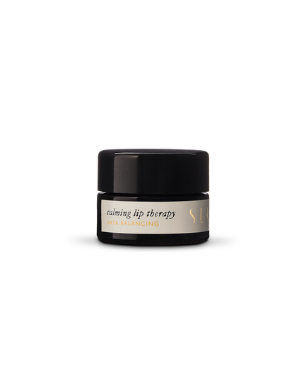 A small, opaque black jar with a screw-on lid contains Surya's "Calming Lip Therapy," subtitled "Vata Balancing." The container features a simple, minimalist design with gold accents. This moisturizing treatment boasts a creamy formula made from organic ingredients.