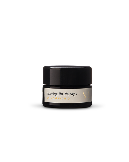 A small, opaque black jar with a screw-on lid contains Surya's "Calming Lip Therapy," subtitled "Vata Balancing." The container features a simple, minimalist design with gold accents. This moisturizing treatment boasts a creamy formula made from organic ingredients.