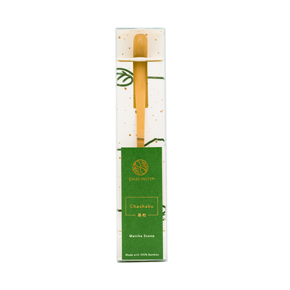 A bamboo chashaku (matcha scoop) is neatly packaged in a white and green box with gold speckles, perfect for matcha preparation. The green label on the box reads "Chafinity Tea Chashaku Matcha Bamboo Scoop Made with 100% Bamboo" along with a logo.