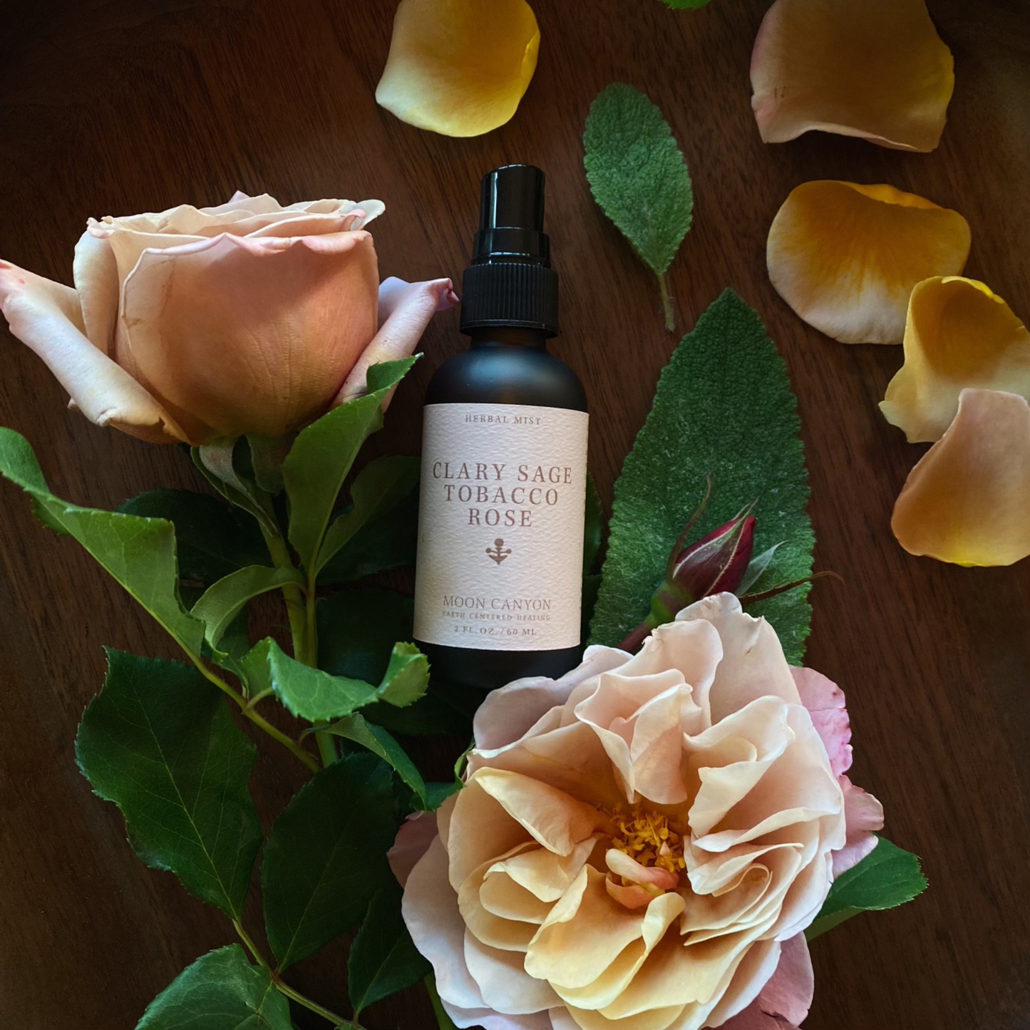 A spray bottle labeled "Clary Sage, Rose & Tobacco Herbal Mist" by Moon Canyon Healing lies amidst a display of pink roses and green leaves on a dark wooden surface. Loose yellow rose petals are scattered around, creating an elegant and natural scene. This energetic mist, crafted from organic ingredients, embodies both sophistication and groundedness.