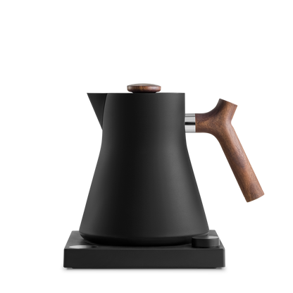 The Corvo EKG Electric Kettle by Fellow Wholesale is displayed. This sleek black kettle features a modern conical design and is precision engineered with variable temperature control. It includes a stylish wooden handle and knob on the lid, all while sitting on a matching black base for heating.