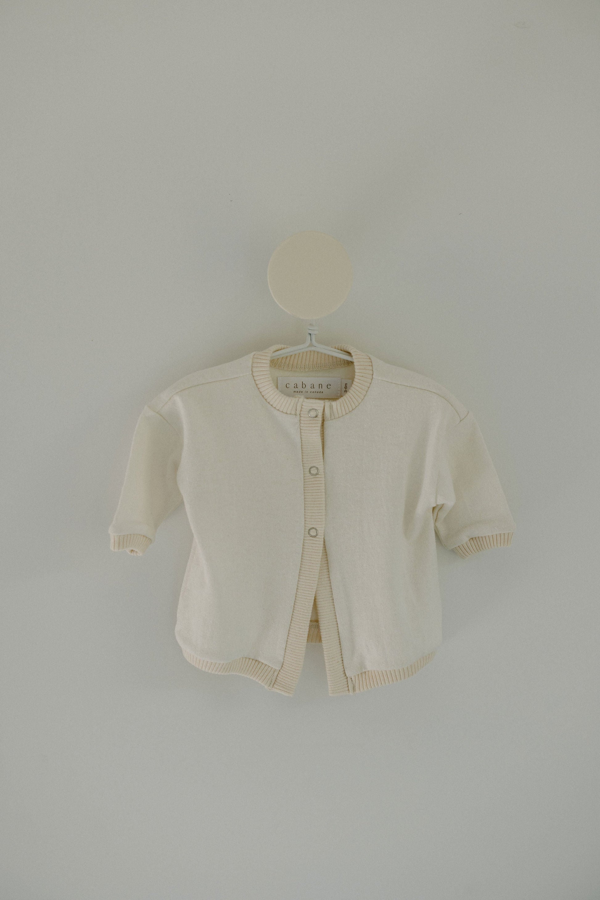 A minimalist and simple Soul Sweater Baby + Kid from cabane childrenswear is displayed on a hanger against a light gray background. It's a small, cream-colored, boxy 3-snap cardigan made from organic cotton and hemp, featuring ribbed edges on the cuffs, hem, and collar. The label inside reads "cabane.