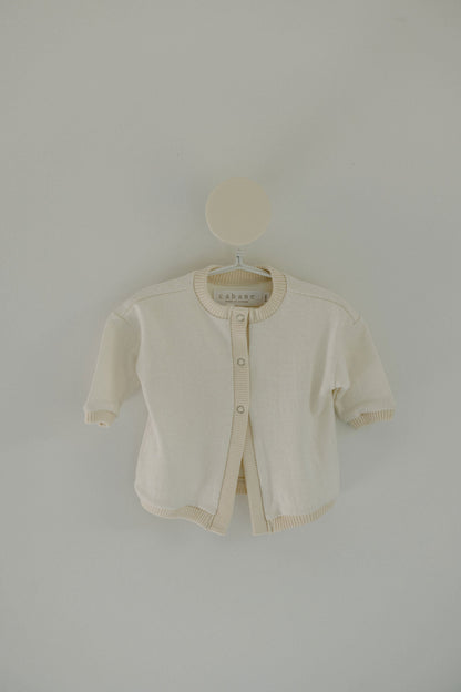 A minimalist and simple Soul Sweater Baby + Kid from cabane childrenswear is displayed on a hanger against a light gray background. It's a small, cream-colored, boxy 3-snap cardigan made from organic cotton and hemp, featuring ribbed edges on the cuffs, hem, and collar. The label inside reads "cabane.