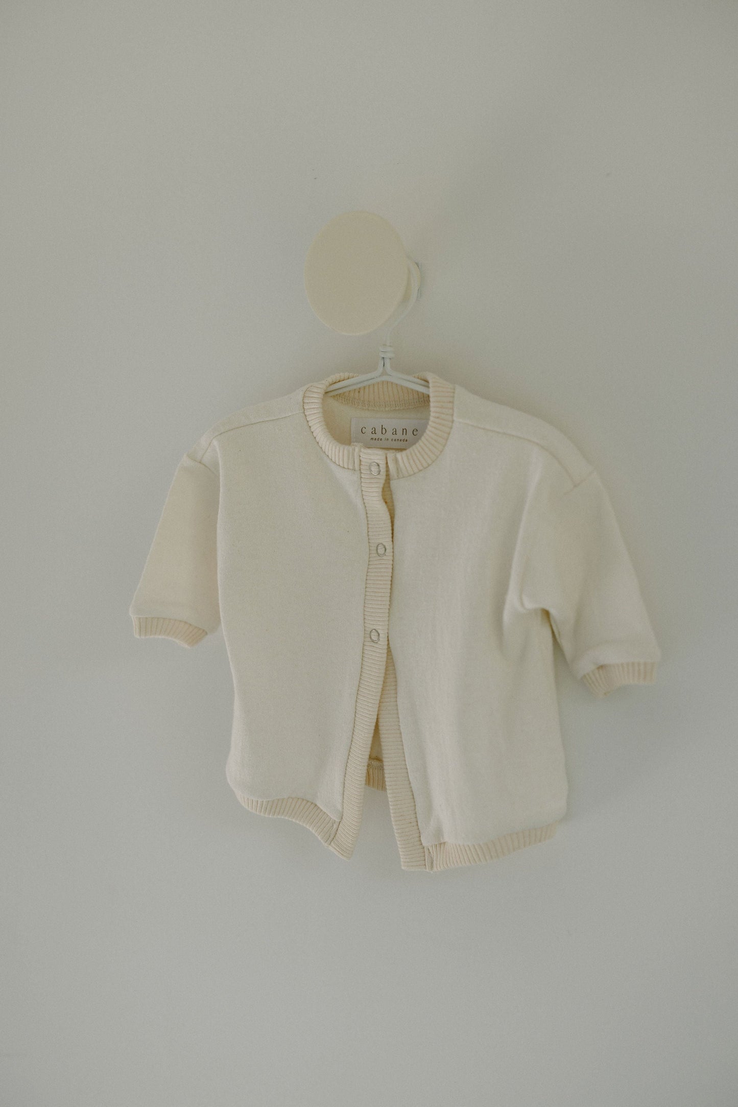 A small, off-white Soul Sweater Baby + Kid with beige trim is hanging on a circular peg against a light-colored wall. The boxy, 3-snap cardigan made from organic cotton and hemp features long sleeves and buttons down the front. The label inside the collar reads "cabane childrenswear.