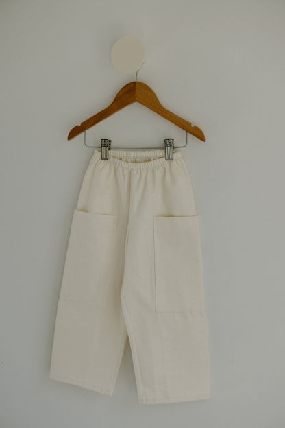 The Pantalon D'Artiste in Hemp Twill by cabane childrenswear, featuring a cream-colored flexible fit and large pockets, hangs on a wooden hanger against a plain light background. The pants have an elastic waistband, and the hanger is attached to a round wall hook.