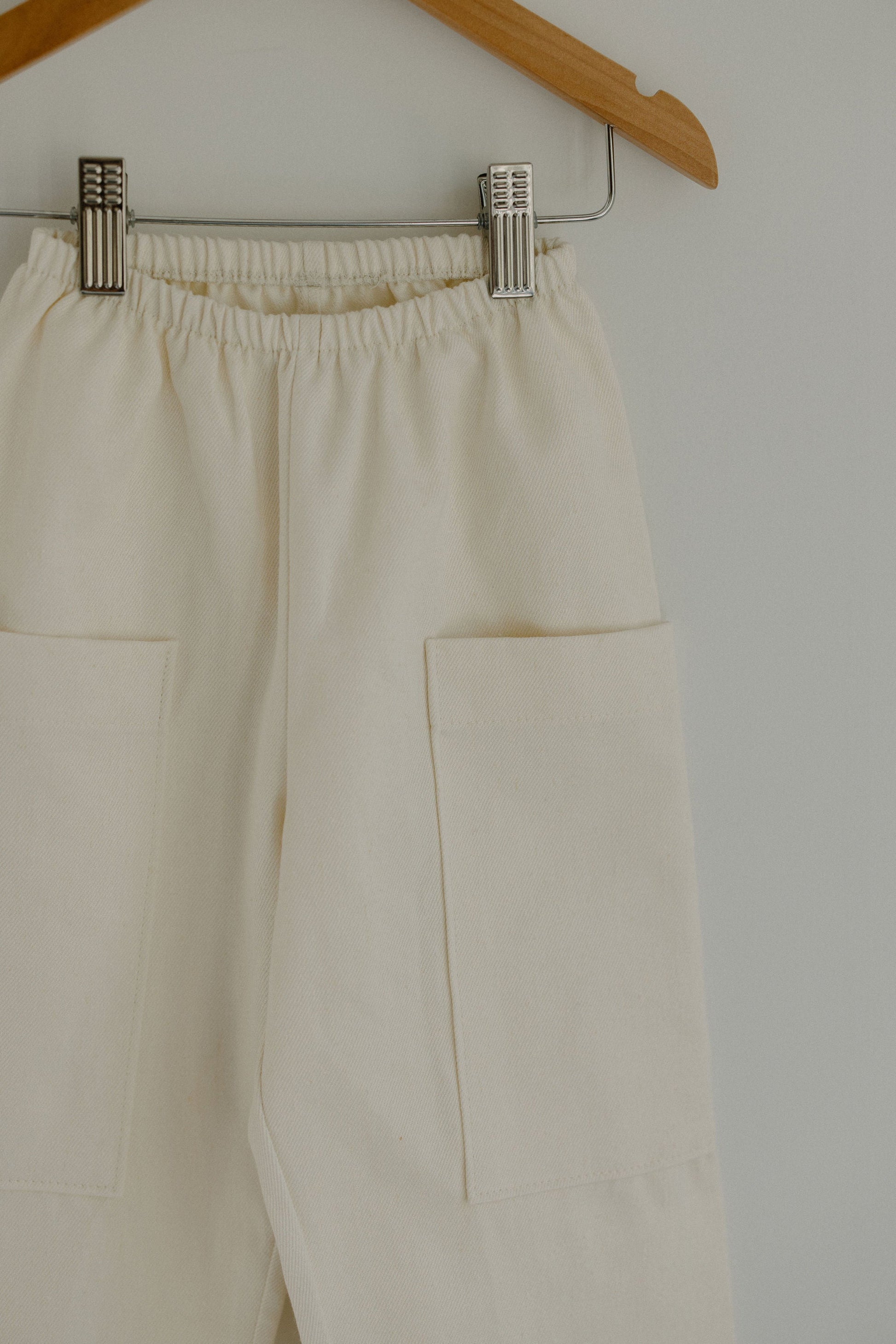 The Pantalon D'Artiste in Hemp Twill by cabane childrenswear hangs on a wooden hanger against a light background. These handcrafted children's pants, made in a beige flexible fit, feature an elastic waistband and large front pockets, secured with metal clips.