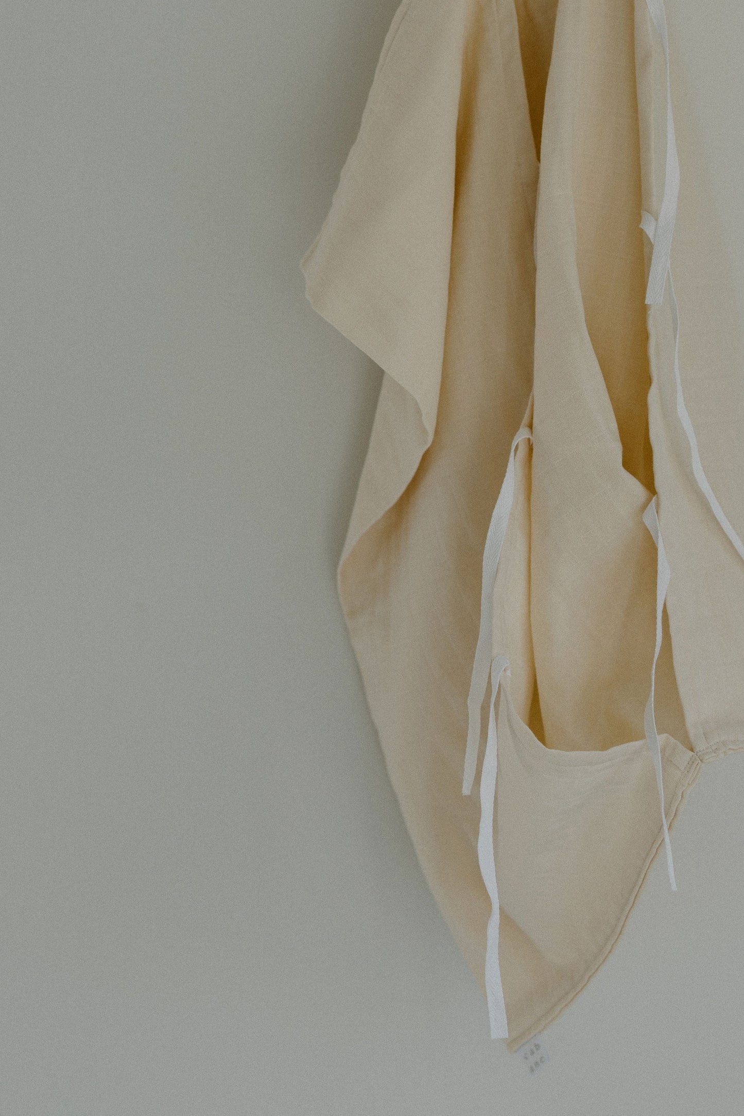 A beige organic cotton fabric with white ties from the cabane childrenswear Organic Garment Bag collection hangs against a plain, light-colored wall. Some edges of the fabric appear folded and draped, creating soft shadows and gentle folds, reminiscent of children's clothing or a well-loved lay-flat bag.