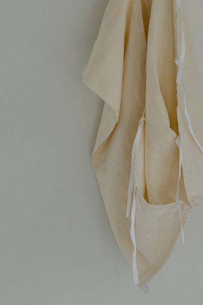 A beige organic cotton fabric with white ties from the cabane childrenswear Organic Garment Bag collection hangs against a plain, light-colored wall. Some edges of the fabric appear folded and draped, creating soft shadows and gentle folds, reminiscent of children's clothing or a well-loved lay-flat bag.