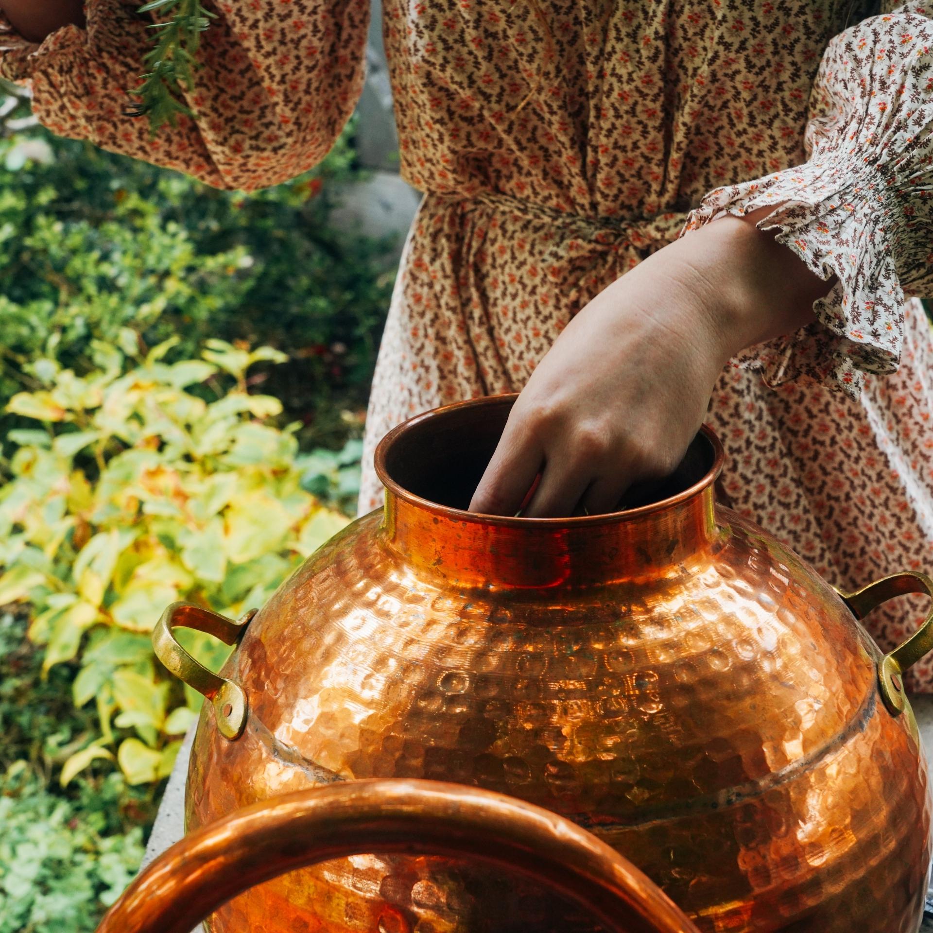 A person in a floral dress is placing their hand inside a large, hammered copper pot outdoors, surrounded by greenery and plants. They are using organic ingredients to prepare the Herbal Skin Toner by Moon Canyon Healing.