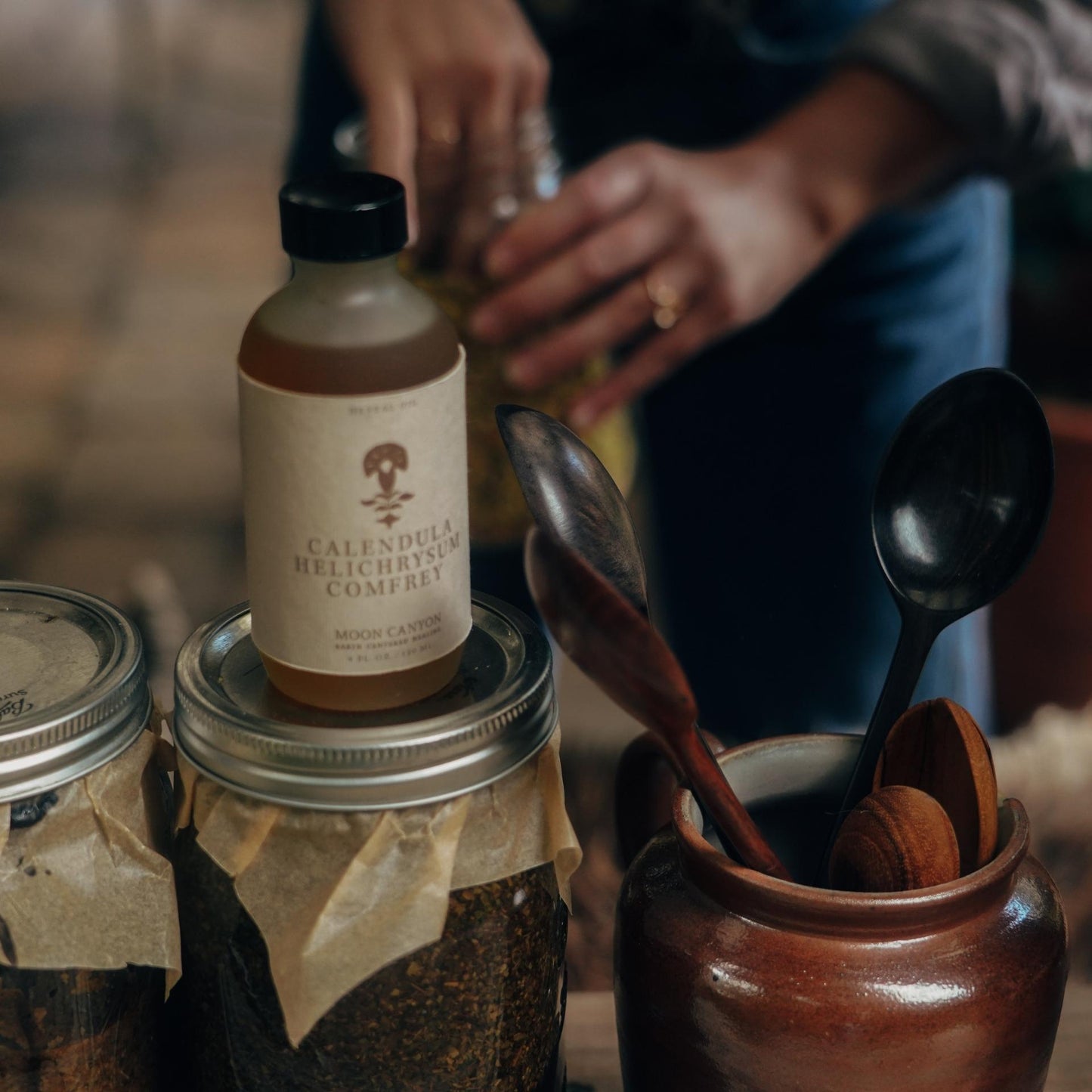 A close-up of jars containing herbal products. One jar, labeled "Calendula, Helichrysum & Comfrey Herbal Oil" by Moon Canyon Healing, stands out. In the foreground, a ceramic container holds wooden spoons. Blurred hands are seen in the background working with another jar, possibly creating a soothing face and body oil.