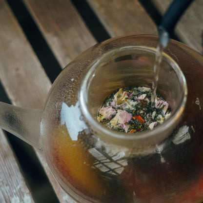 A close-up view from above shows hot water being poured into a glass teapot containing Moon Canyon Healing's Holy Heart Tea Blend. Inside the teapot, you can see dried flowers and loose leaf tea infused with adaptogenic herbs. The teapot rests on a wooden surface, creating a serene moment of heart-centered awareness.