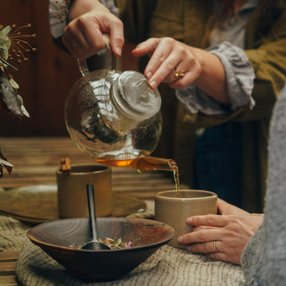 A person pours Moon Canyon Healing's Holy Heart Tea Blend from a glass teapot into a beige mug held by another person. A wooden bowl with a spoon inside and a rustic tablecloth adorn the foreground, creating a warm, cozy atmosphere filled with natural elements and heart-centered awareness.