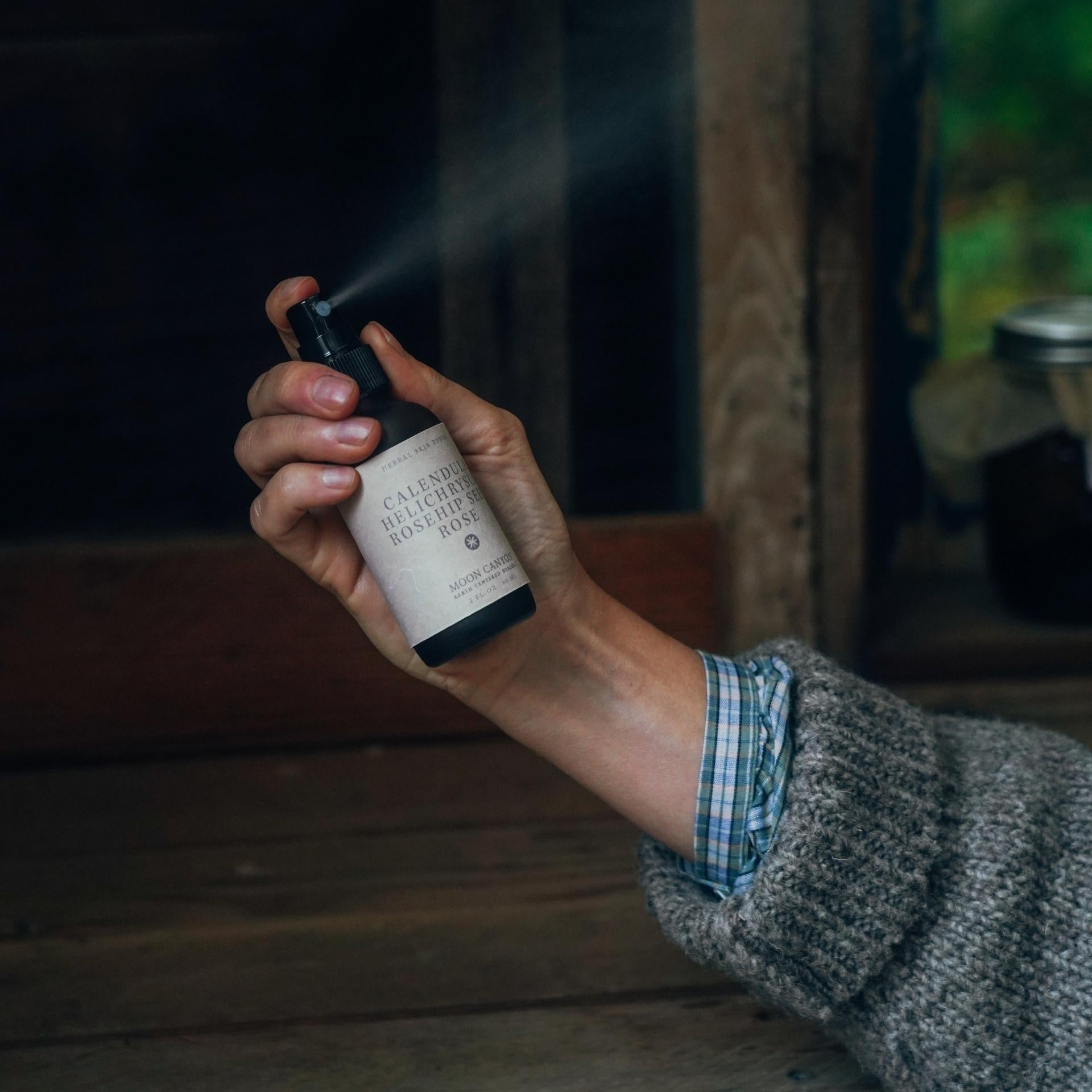 A hand, clad in a gray knitted sleeve and checkered shirt, is holding and spraying a bottle labeled "Herbal Skin Toner" from Moon Canyon Healing. Made with organic ingredients, the background exudes a rustic charm with wooden elements and a glimpse of a jar on a shelf.