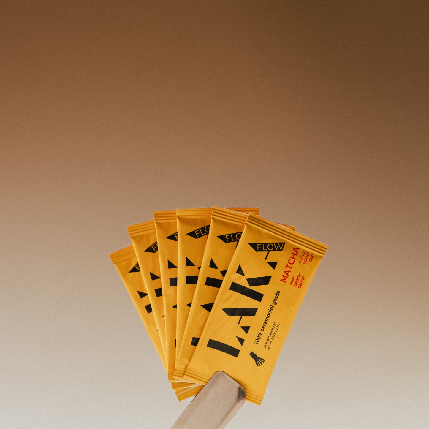 A fan-shaped arrangement of five yellow LAKA Matcha™ Sticks, held together by a white hand at the bottom left against a gradient brown background. The packaging features minimal black text and a minimalist design, highlighting its ceremonial-grade matcha for energy and metabolism from the brand LAKA.