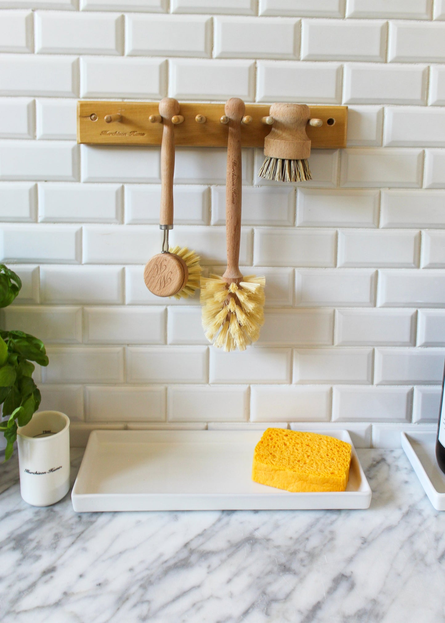 The All-Brush Bundle from Murchison-Hume holds three wooden dish brushes with natural fiber bristles hanging on hooks against a white subway tile backsplash. Below them, a white tray on a marble countertop contains a yellow sponge. A small plant and a white mug are nearby, adding charm to this eco-friendly setup.