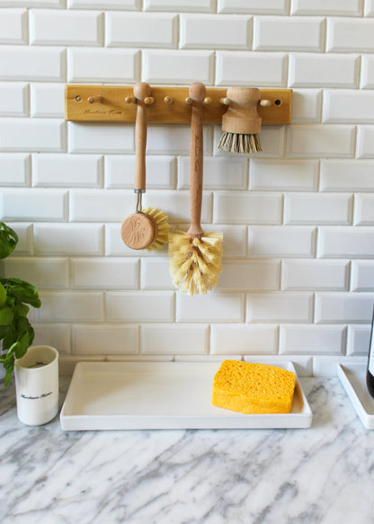 The All-Brush Bundle from Murchison-Hume holds three wooden dish brushes with natural fiber bristles hanging on hooks against a white subway tile backsplash. Below them, a white tray on a marble countertop contains a yellow sponge. A small plant and a white mug are nearby, adding charm to this eco-friendly setup.