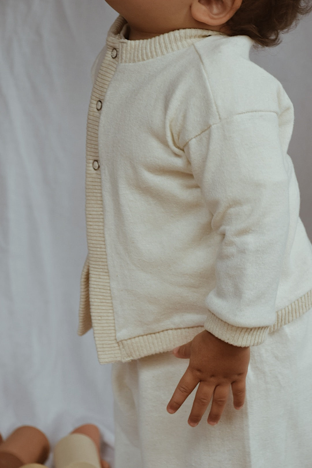 A toddler wearing a cream-colored, boxy Soul Sweater Baby + Kid and matching pants by cabane childrenswear, both crafted from organic cotton and hemp, stands against a neutral background. Only the upper body and one arm of the child are visible in the image. The child is looking slightly upwards, with their head mostly out of the frame.