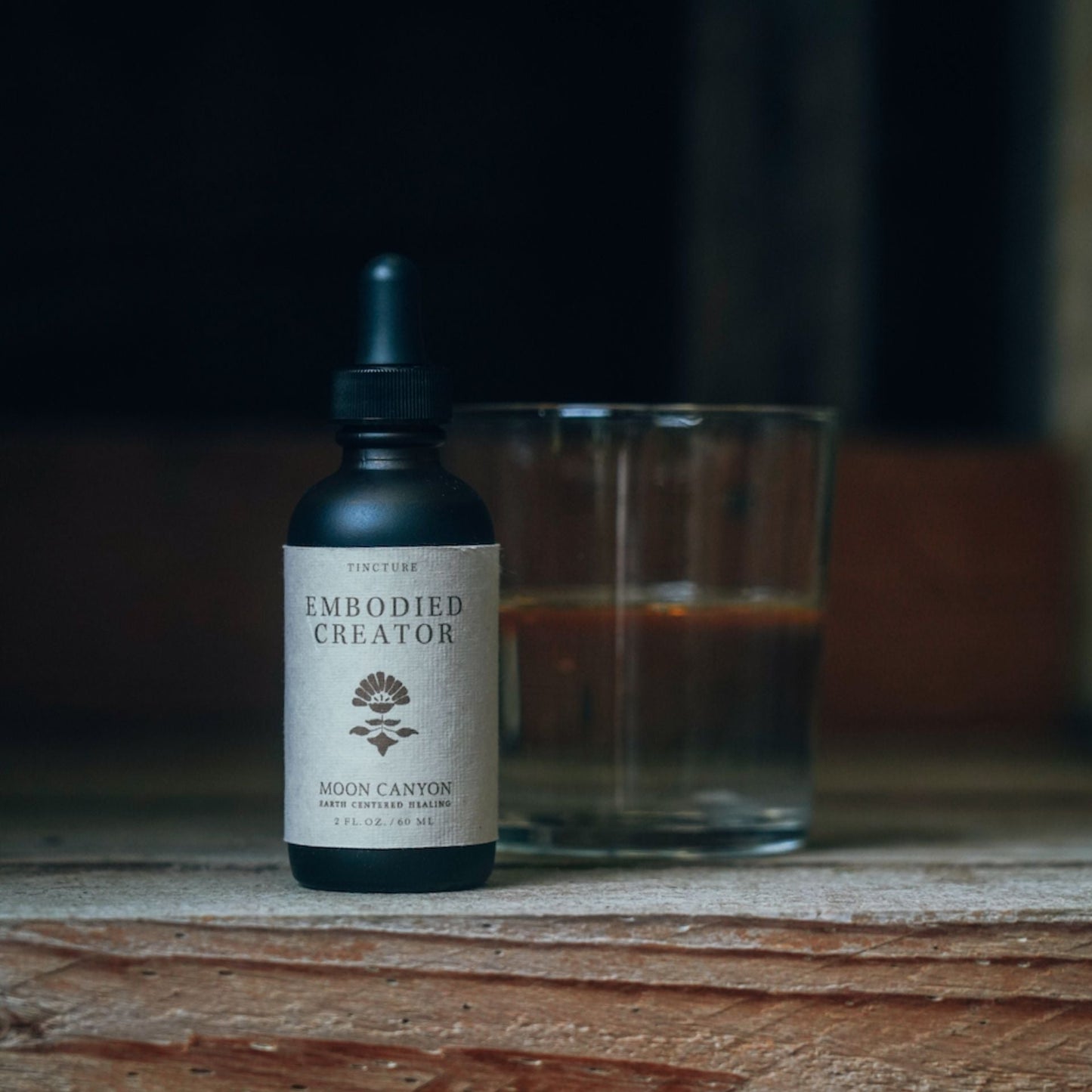 A small dark bottle labeled "Embodied Creator Tincture" by Moon Canyon Healing, equipped with a dropper top, sits on a rustic wooden surface. The bottle rests beside a half-filled transparent glass, with the background softly blurred to spotlight the adaptogenic herbs within.