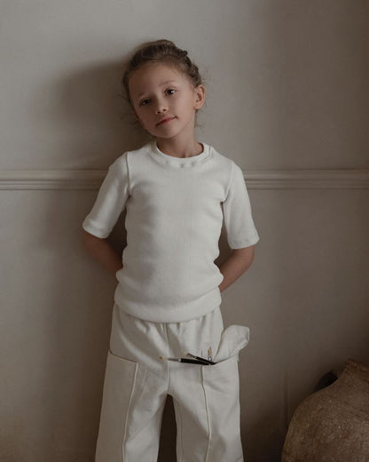 A child wearing a white shirt and the Pantalon D'Artiste in Hemp Twill by cabane childrenswear stands against a wall, hands behind their back, with a small paintbrush tucked in a pocket. The room is softly lit, creating a calm atmosphere.