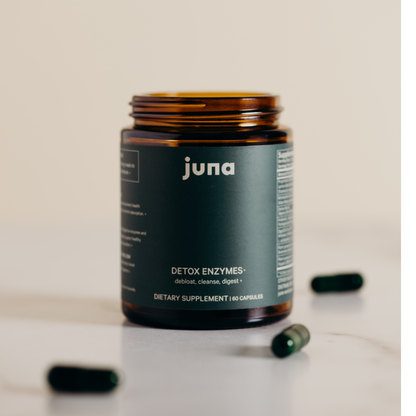 A dark amber jar labeled "Juna Detox Digestive Enzymes" rests on a smooth surface, surrounded by scattered green capsules. Promising to "debloat, cleanse, and digest," this potent blend of digestive enzymes and spore-based probiotics offers 60 dietary supplement capsules for optimal gut health.