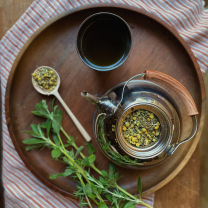 A top view of a wooden tray on a striped cloth, showcasing a metal teapot filled with Moon Canyon Healing's Gut Knowing Tea Blend, a black cup brimming with tea, a wooden spoon containing dried herbs from the blend, and fresh herbs lying next to them. The scene exudes a cozy, rustic ambiance perfect for mood support.